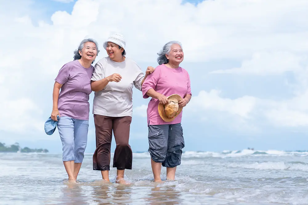Three elderly women smile and walk together in shallow water at the beach on a bright day, enjoying each others company.