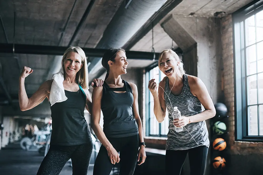 Three women in workout clothes smile and laugh together in a gym. One flexes her arm, another holds a water bottle, and gym equipment and windows are visible in the background.