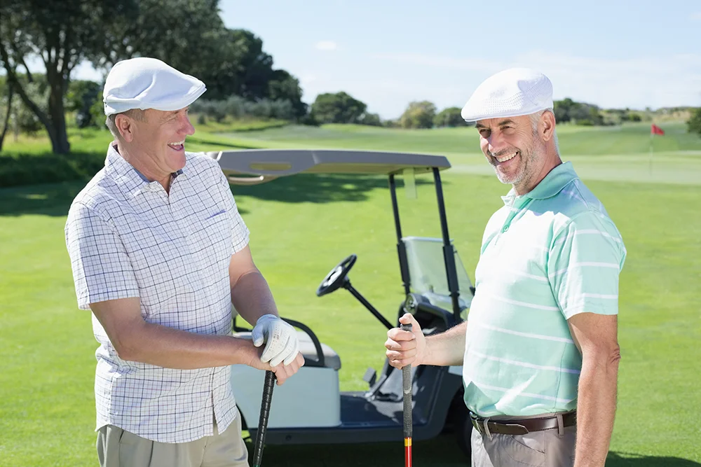 Two older men in golf attire stand on a golf course, smiling and holding clubs, with a golf cart and green grass in the background.