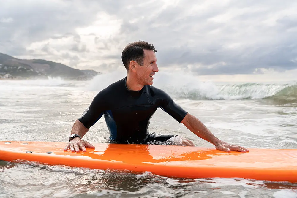 A man in a wetsuit sits on an orange surfboard in the ocean, smiling and looking to the side as waves roll in behind him under a cloudy sky.