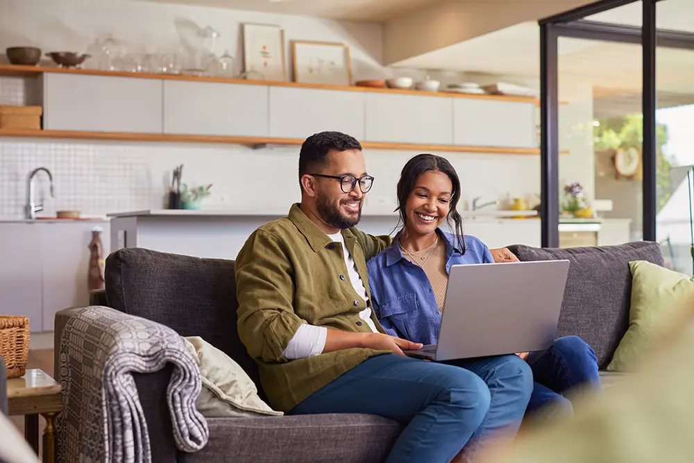 A smiling couple sits on a gray couch in a modern living room, looking at a laptop together. The kitchen is visible in the background, and sunlight streams in from large glass doors.