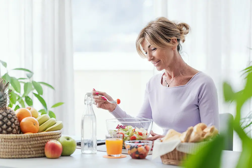 A woman sits at a table, smiling while eating salad. The table holds a fruit basket, a glass of orange juice, a bread basket, a bowl of fruit salad, and a water bottle. Bright natural light fills the room.